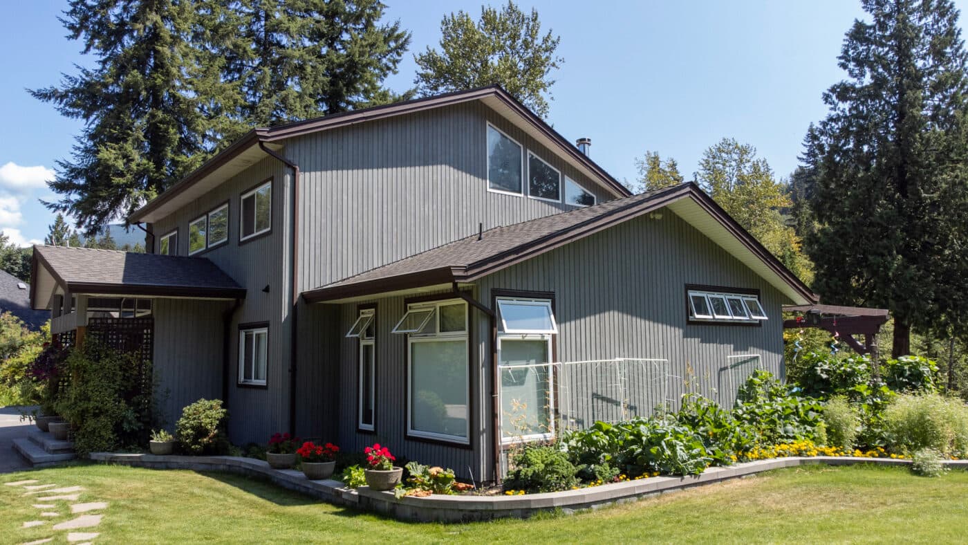 Side elevation of a grey wood-sided home with white windows and garden beds